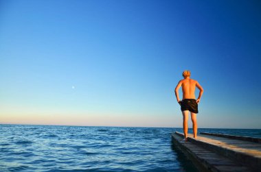 Tek başına duran ve denize bakan genç adam. Yaz akşamından kalma duvar kâğıdı ve montajın için yer var.