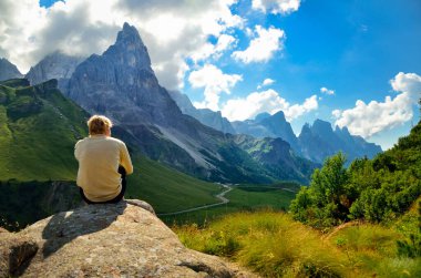 Genç adam yürüyüş sonrası kayanın üzerinde dinleniyor, İtalyan Dolomite tepesinin altındaki yeşil dağlara bakıyor..