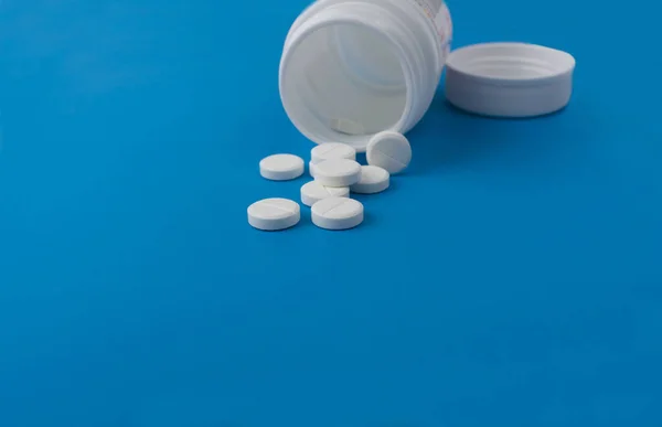 Kapaklı plastik bir kavanozda beyaz tabletler, izole edilmiş mavi bir arkaplanda yatıyor.
