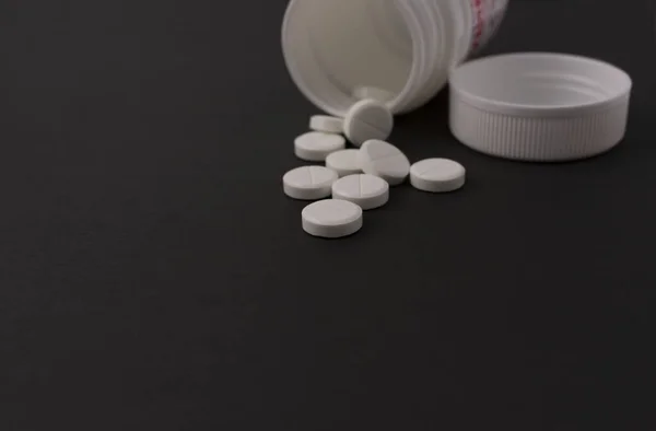 Kapaklı plastik bir kavanozdaki beyaz tabletler izole edilmiş siyah bir arkaplanda yatıyor.