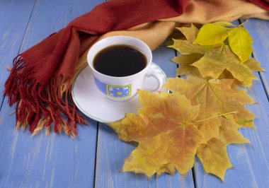 Sarı yapraklar ve sıcak bir eşarbın yanında, ahşap mavi arka planda bir kahve fincanı.
