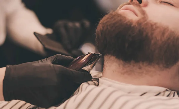 Bearded male sitting in an armchair in a barber shop while hairdresser shaves his beard