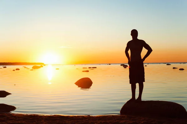 Młody, sportowa sylwetka stojący nad brzegiem morza na zachód słońca — Zdjęcie stockowe