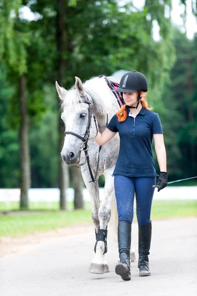 Young teenage redhead girl leading gray horse from training — Stock Photo, Image