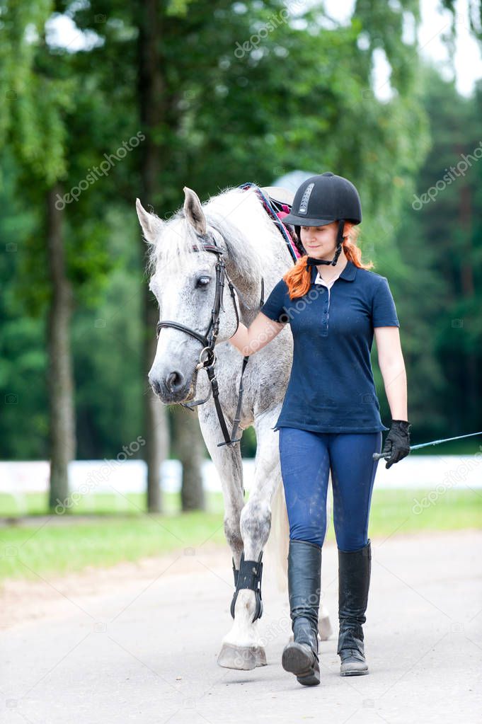 Young teenage redhead girl leading gray horse from training