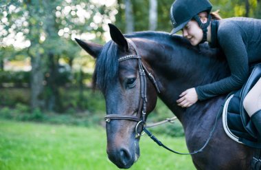Binici kadın ata biniyor ve kestane rengi at boynunu okşuyor. Açık havada canlı renkli, filtreli yatay yaz dönemi görüntüsü