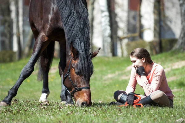 若い十代の少女の所有者の近くに彼女の栗の馬に緑の草の上に座っている 活気に満ちた多色の春屋外水平画像 ストックフォト