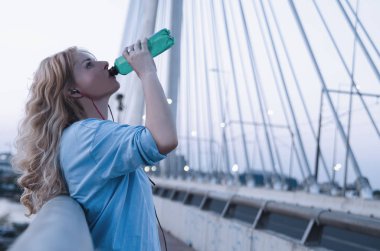 Şehirde egzersiz yaparken plastik şişeden su içen bir kadın. Sarışın, güzel bir kadın enerji içeceği içiyor ve koşuya ara veriyor.