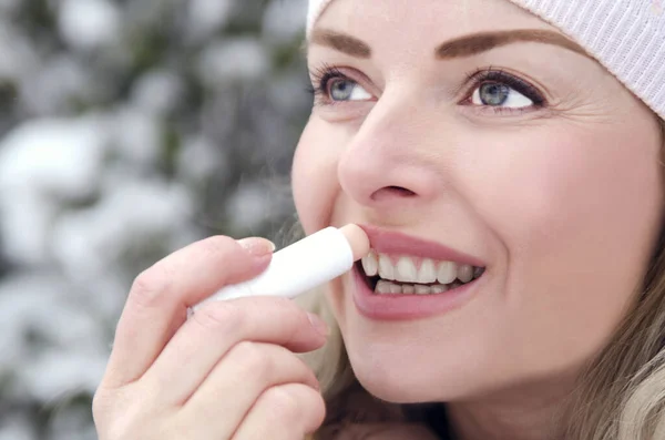 Pretty girl applying lip balm on cold winter day outdoor. Nordic woman in warm clothes putting cosmetic protection for lips on snowy season