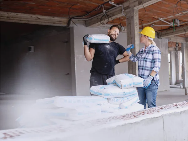 İnşaat alanında inşaat işçisi ve kadın mühendis çimento çantası taşıyor. Kadın ve erkek yeni ev inşaatında birlikte çalışıyor.