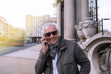 Şehir sokaklarında akıllı telefon kullanan şık, yaşlı ve yakışıklı bir adam. Mutlu yaşlı, yakışıklı, gri saçlı adam cep telefonuyla konuşuyor.
