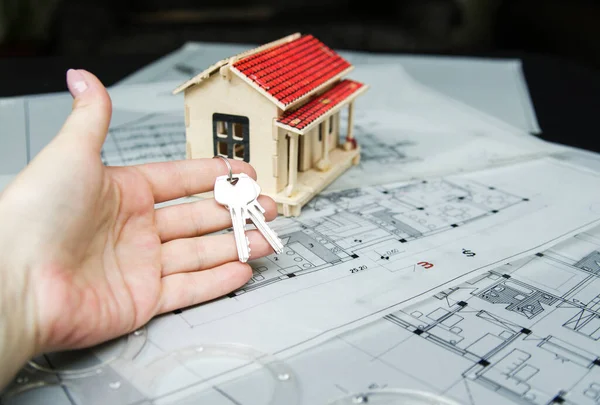 Yeni evin anahtarlarını taşıyan kadın parmağı. Mimari proje üzerine ev modeli. Yeni evin anahtarlarıyla mavi baskı. Boş bir ev için yatırım. Bankaya mevduat kredisi.