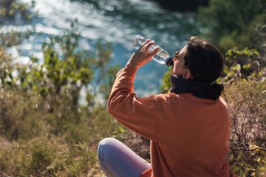 Güneş gözlüklü ve turuncu kazaklı kadın içme suyu içiyor.