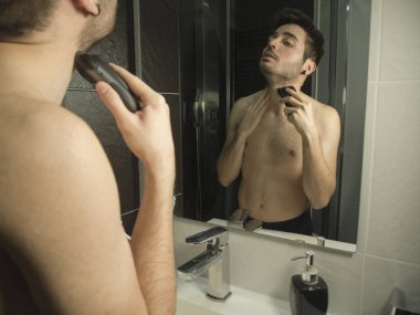 Portrait of handsome caucasian teenager reflection in from of the mirror, shaving with a shaver