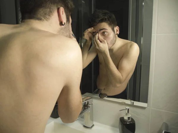 Portrait of handsome caucasian teenager reflection in from of the mirror, waxing his eyebrows with tweezers