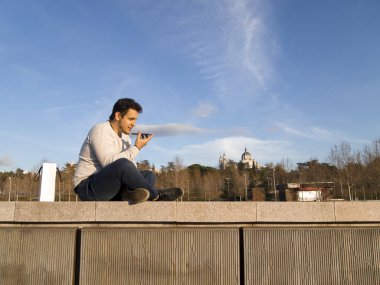 Genç beyaz adam telefonuna ses kaydediyor. Madrid Rio 'da oturuyor. Arkasında da Almudena Katedrali var.