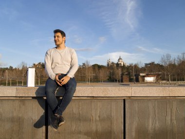 Young handsome caucasian man smiling, with the phone in his hands, seated at Madrid Rio with the Almudena Cathedral behind him
