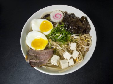 Ramen: Asian noodle soup with beef, eggs, fungi, vegetables, tofu, seaweed and naruto in a bowl. Black background. Top view.