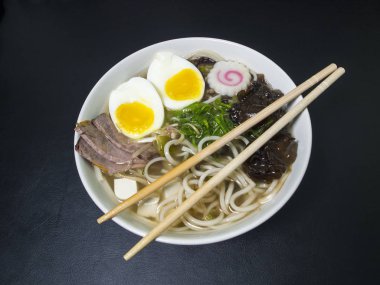 Ramen: Asian noodle soup with beef, eggs, fungi, vegetables, tofu, seaweed and naruto with chopsticks on the bowl. Black background. Top view.