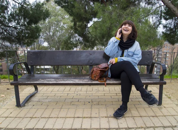 Portrait of a urban young stylish smiling woman in bright modern clothes sitting on a bench. She is laughing and talking on the smartphone. Lifestyle and urban concept.