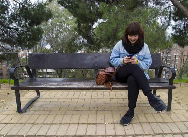 Portrait of a urban young stylish smiling woman in bright modern clothes sitting on a bench. She is writing on her smartphone. Lifestyle and urban concept.