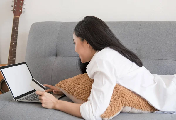 Beyaz elbiseli güzel bir kadın, kanepede dizüstü bilgisayarla oynuyor. İş fikirleri ve online alışveriş.
