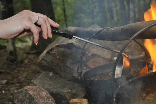 Female hand with a wedding ring and a ladle of soup. The soup is already in the pot And the pot hangs on a stick over the fire.