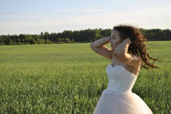 Bride at a photo shoot in the field. The bride waves her hair in the wind. A bride in a white dress looks into the distance. Forest in the background.