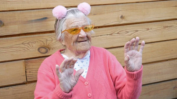 Grandma in a pink blouse, orange glasses and pink ears on the rim, waving happily. In the background is a wooden background.