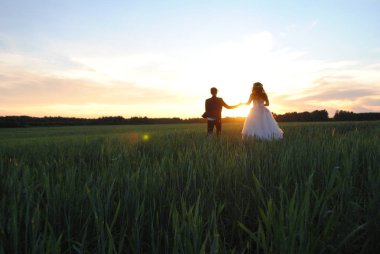 Yeni evliler güneşe doğru el ele tutuşarak buğday tarlasında koşar..