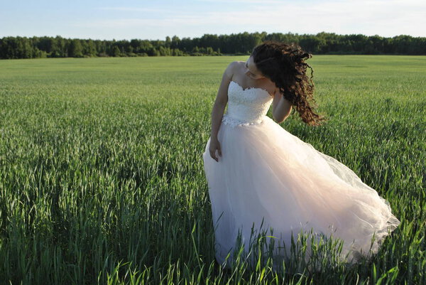 Bride at a photo shoot in the field. The bride with her head down straightens her curly hair. The forest is in the background.