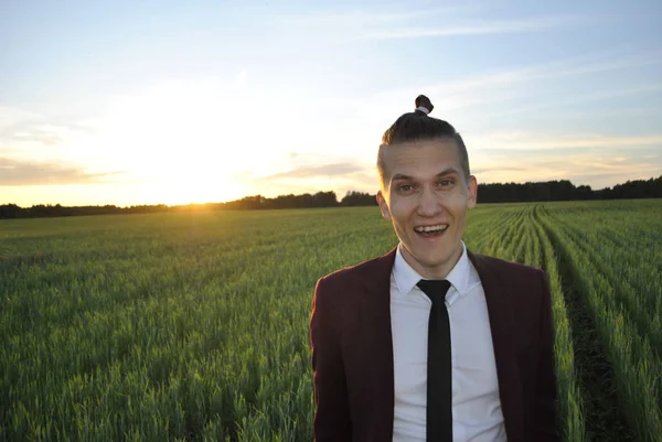 A young guy laughs at the camera in a wheat field. The guy is wearing a maroon suit. An orange sunset shines into the camera.