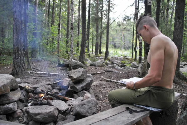 A young reader reads a book in nature.A young guy is sitting by the fire. The fire is burning a little. Fire smoke is spreading in the air.