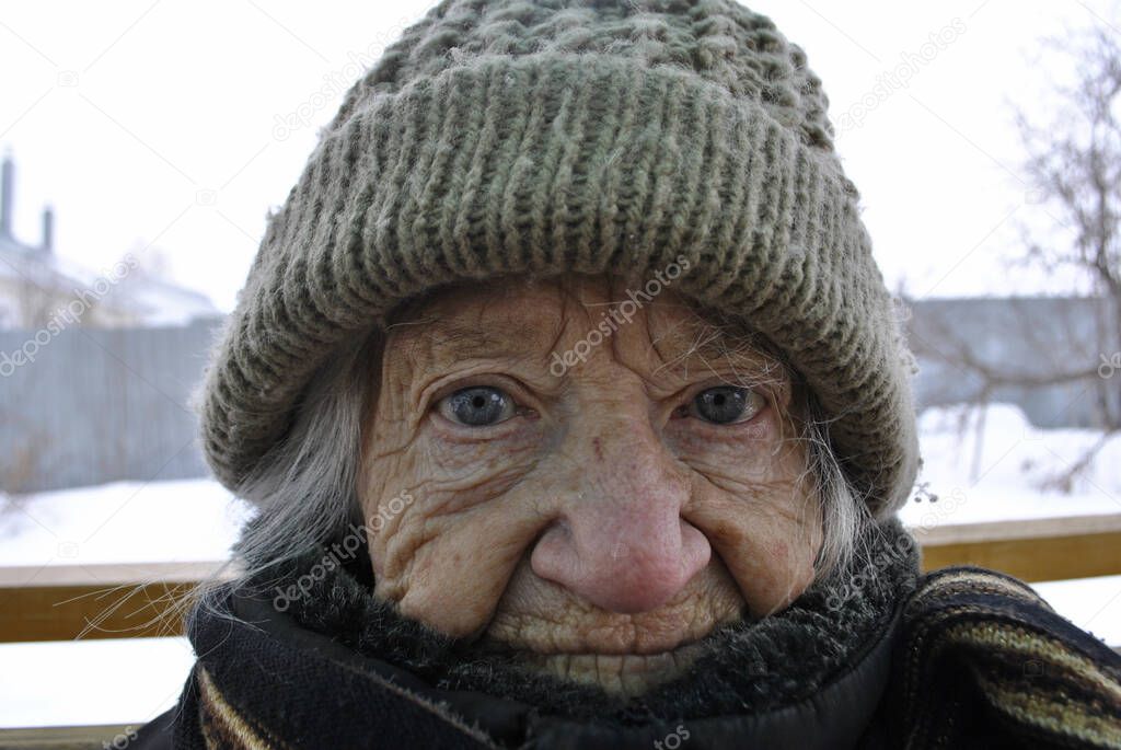 An elderly grandmother looks at the camera with a tired expression. Granny close-up. Grandma is dressed in warm clothes. She wears a hat and scarf.