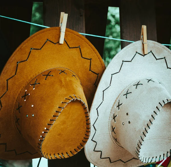 Two decorative leather hats in white and brown with stitching hanging on a line with wooden clothes pegs in close up