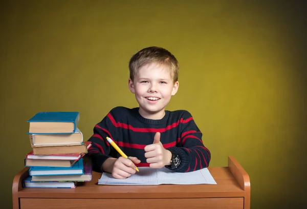 Ragazzo felice che fa i compiti con il pollice alzato, libri sul tavolo . — Foto Stock