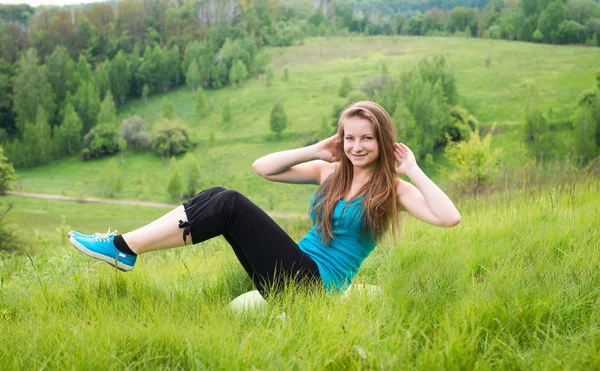 仰卧起坐-健身女孩训练坐在夏天在草上外。快乐合适的女人做侧仰卧起坐与高架腿同时微笑快乐. 免版税图库照片