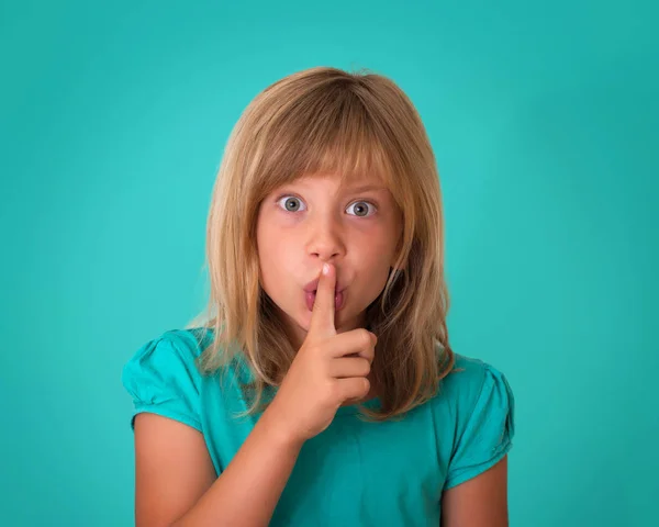 Schönes kleines Mädchen, das den Finger an die Lippen legt und um Stille auf türkisfarbenem Hintergrund bittet. Kind macht bitte leise Geste in Richtung Kamera. — Stockfoto