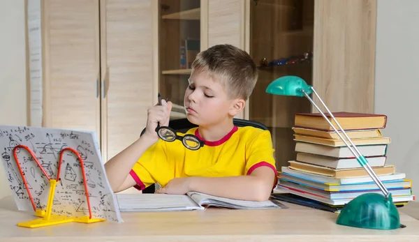 Ragazzo stanco che indossa occhiali divertenti mentre fa i compiti. Bambino con imparare Fotografia Stock