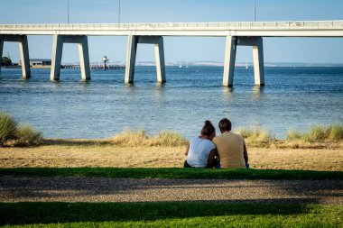 Tatlı bir çift Melbourne 'daki San Remo plajı ve köprüsünün önündeki çimlerde sohbet ediyor.