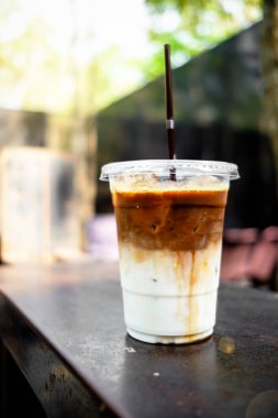 Plastik bardakta servis edilen buzlu karamelli macchiato 'nun yakın plan bir fotoğrafı, paslı metal masaya yerleştirilmiş, kafenin içinde bulanık bir arka planı var.