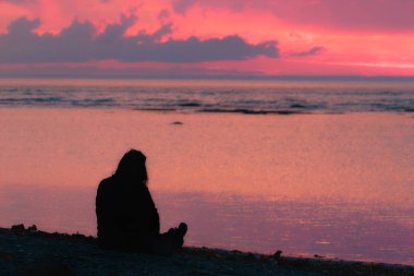 Sahilde oturmuş, gün batımını seyreden bir kadın silueti.