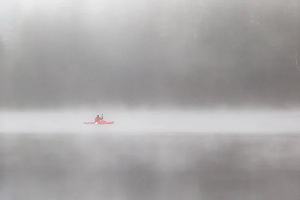 Kayaker in red gear paddling in fog on quiet lake