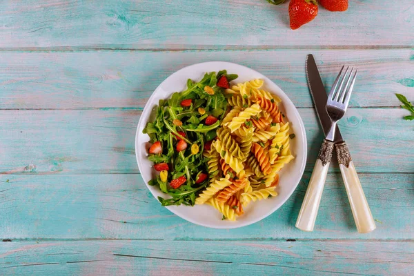 Pasta rotini con rúcula ensalada de fresas con tenedor y cuchillo sobre fondo de madera. Vista superior . — Foto de Stock