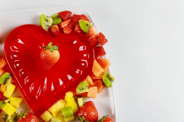 Forma de coração de gelatina vermelha com frutas frescas picadas em um fundo branco . — Fotografia de Stock