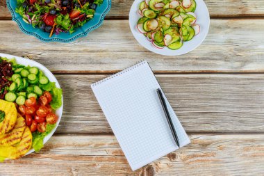 Defter ve kalemle ahşap masada çeşitli salatalar..