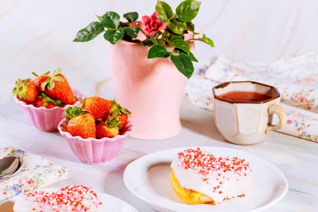 Two sweet heart shaped donut with white glaze and sprinkles on party table with tea, strawberries and rose. Valentine Day concept.