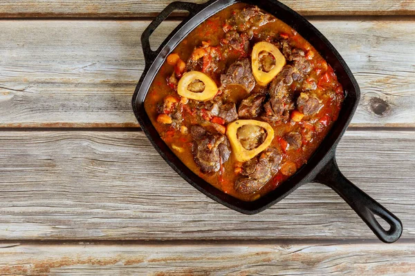 Cast iron pan with ossobuco made from beef shank bone in and vegetable on wooden table.