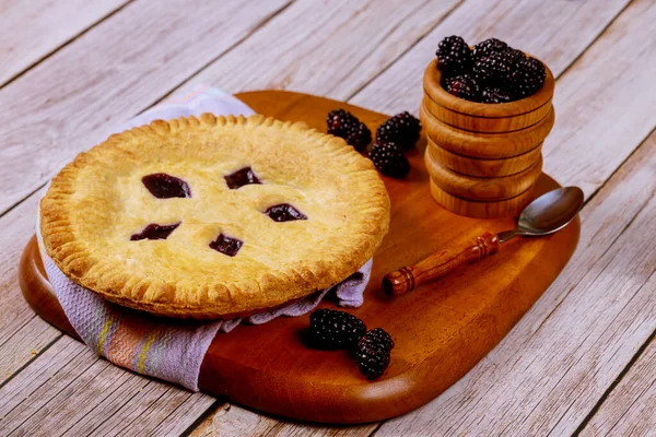 Round blackberry pie on wooden table with spoon and fresh berries.