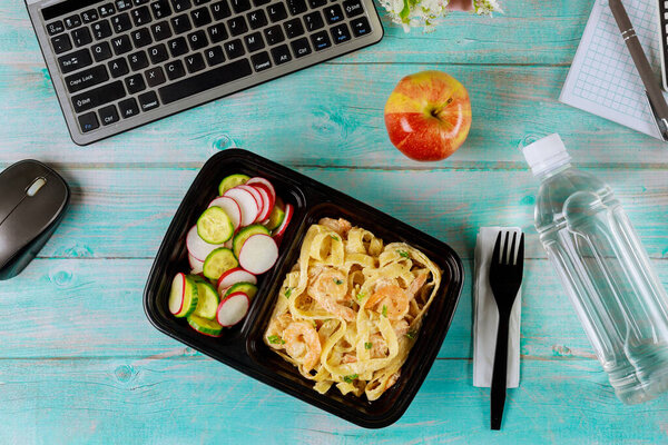 Food delivery to work. Lunch box container with pasta and shrimps, cucumber and radish on wooden table with laptop.